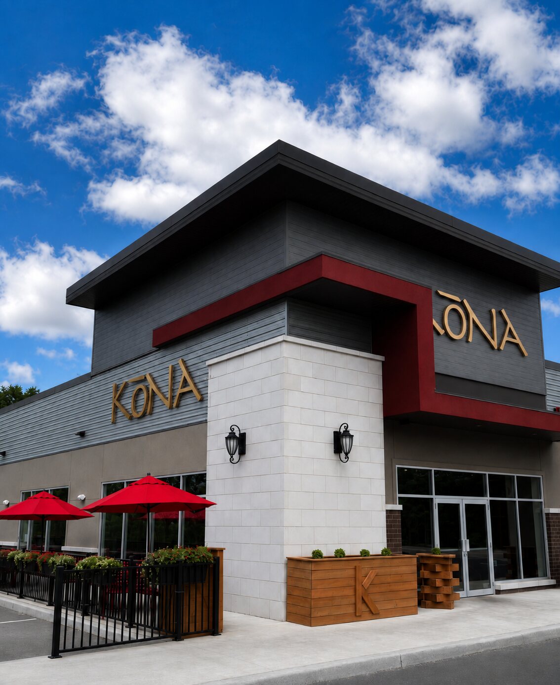 The Kōna Lakeshore storefront — modern grey and red building with gold KŌNA lettering, red patio umbrellas out front and a bright blue sky.