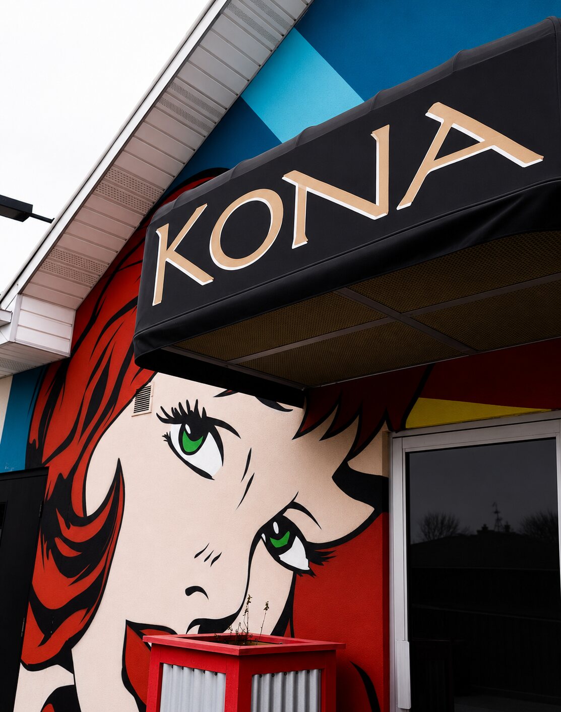 Bold Lichtenstein-style pop-art mural of a red-haired woman on the entrance wall beneath the KŌNA awning.