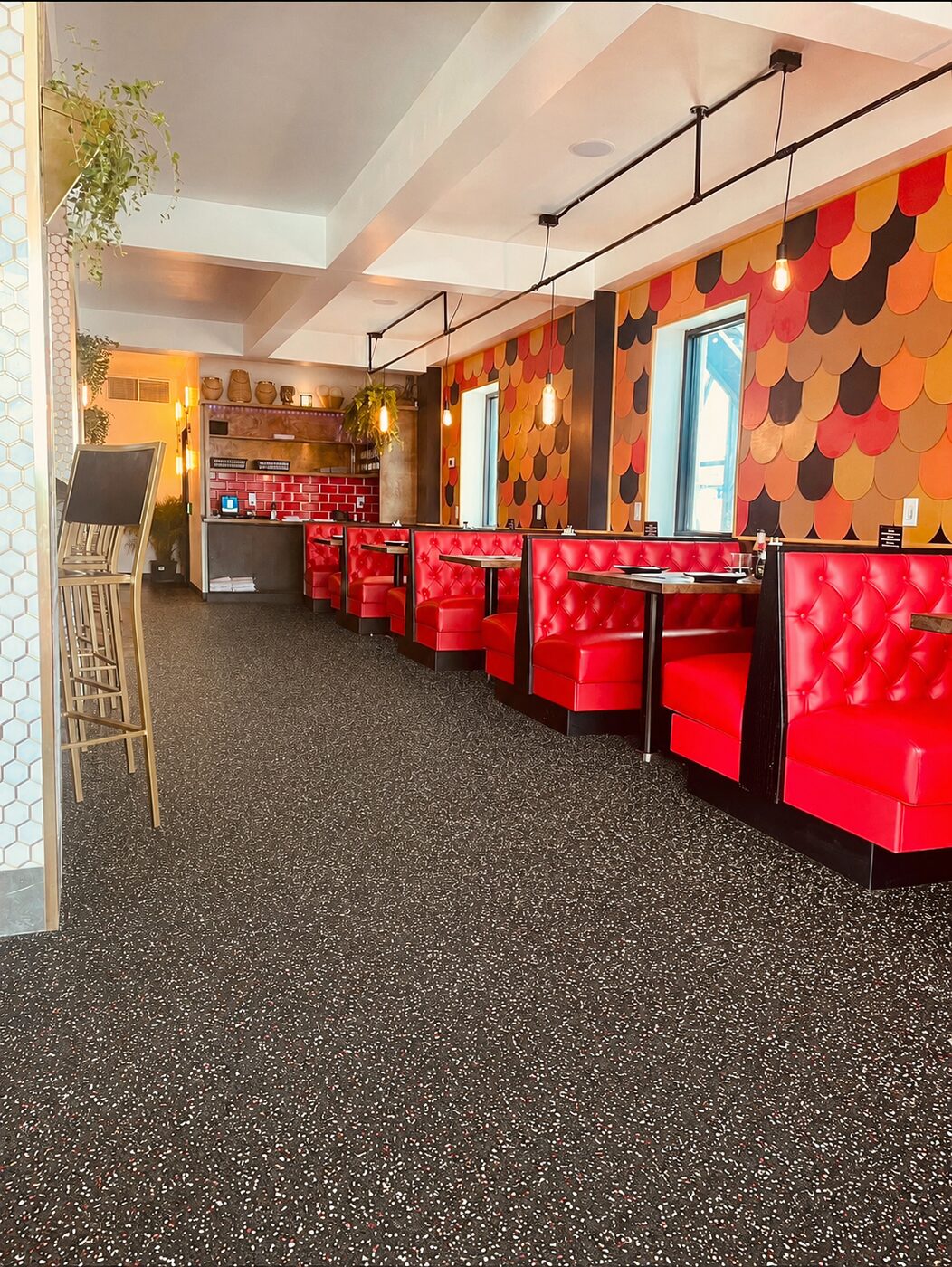 Row of red tufted booths beneath Kōna Tecumseh's scalloped orange, red and gold feature wall, with warm pendant lights and hanging plants.