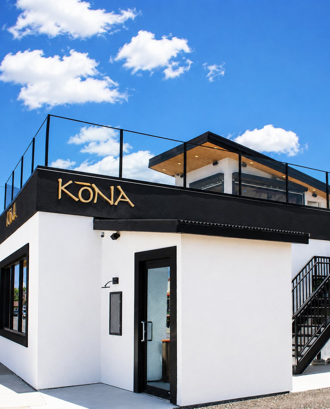 The Kōna Tecumseh storefront — white and black building with the gold KŌNA sign and the rooftop patio railing above, against a bright blue sky.