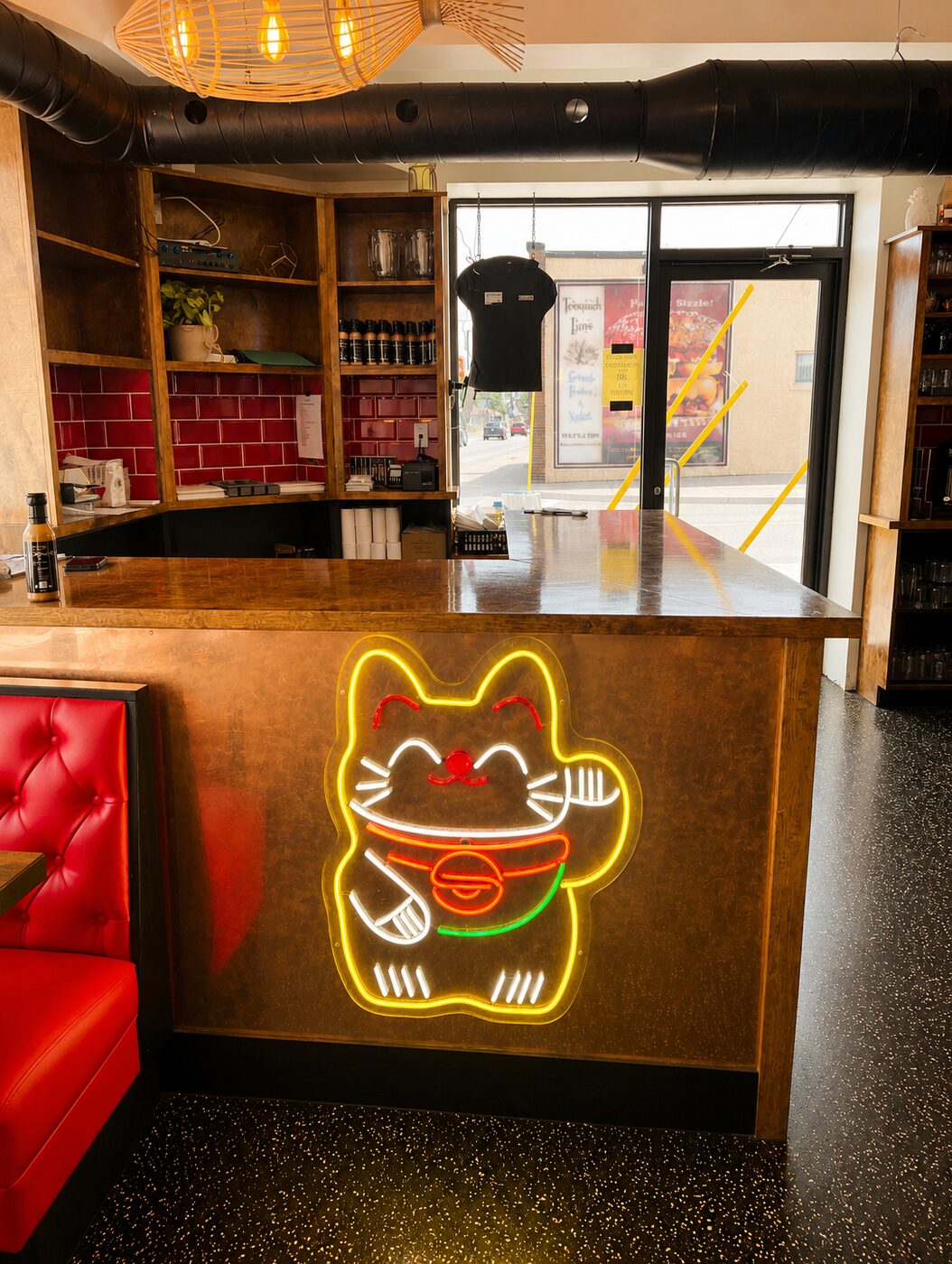 Kōna's signature neon lucky cat glowing gold on the burl-wood host stand, next to a red tufted booth.