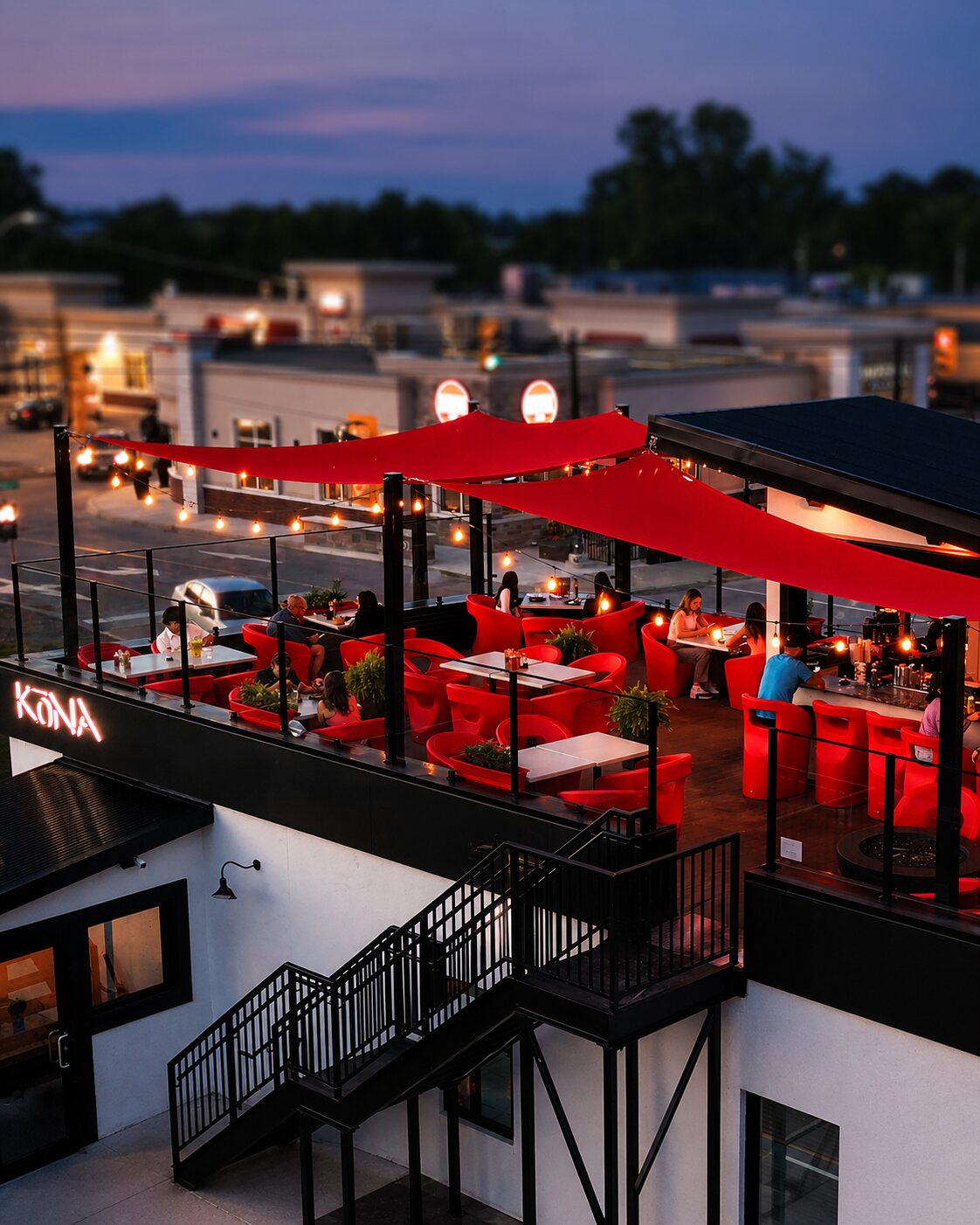 Kōna Tecumseh's rooftop patio at dusk — red sail shades and string lights over red lounge chairs full of guests, the glowing KŌNA neon sign, and the town below under a purple sky.