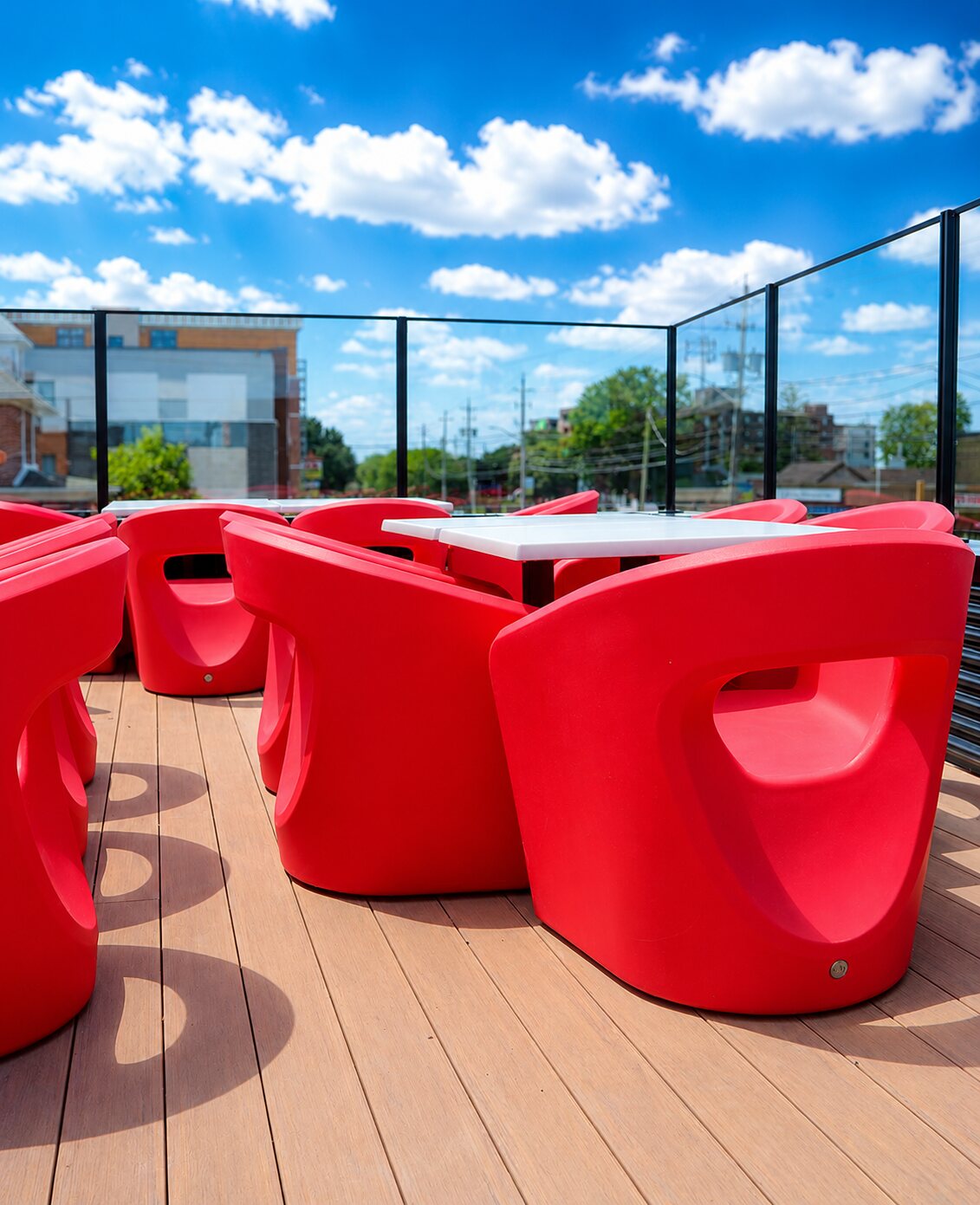 Bold red moulded lounge chairs around white tables on the wood-decked rooftop patio, glass railing and blue sky beyond.