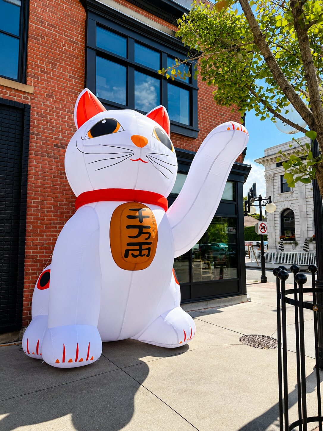 A giant inflatable white lucky cat waving on the sidewalk outside Kōna Walkerville's brick building.