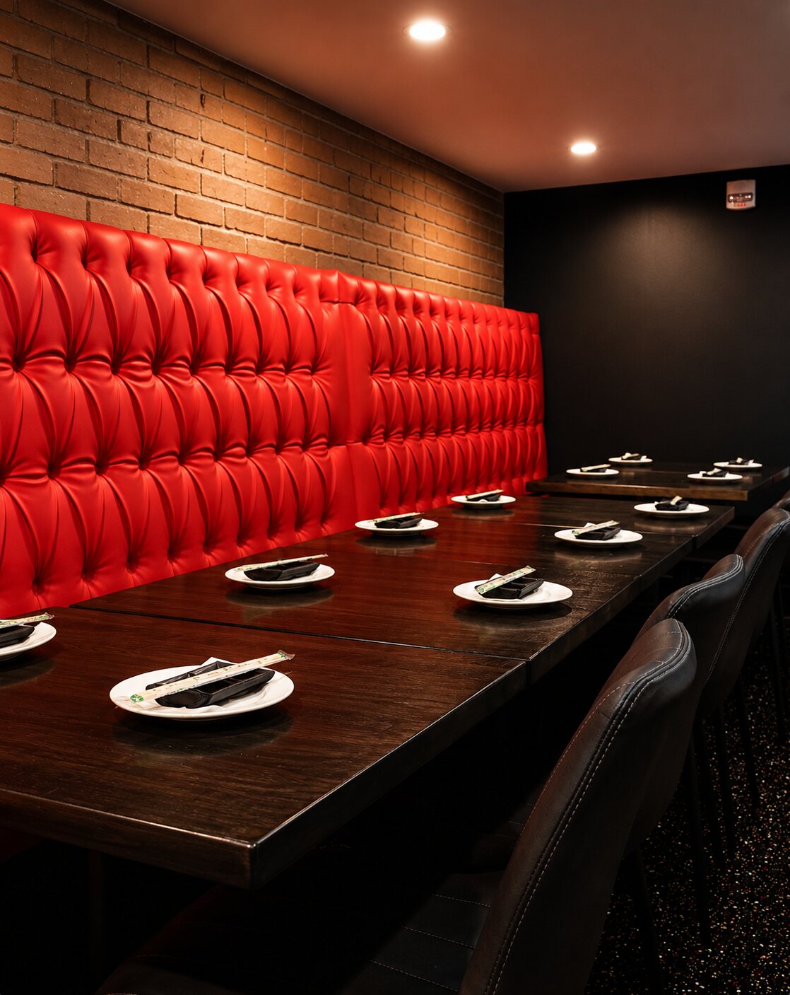 A long private dining table with a big red tufted banquette against exposed brick under moody lighting.